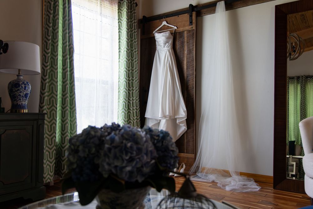 Wedding dress hanging in window light with green curtains and dark furniture in foreground.