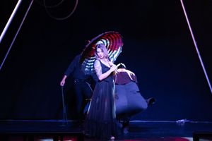 A performer in a flowing dress poses with a parasol under dramatic stage lighting during a theatrical performance.