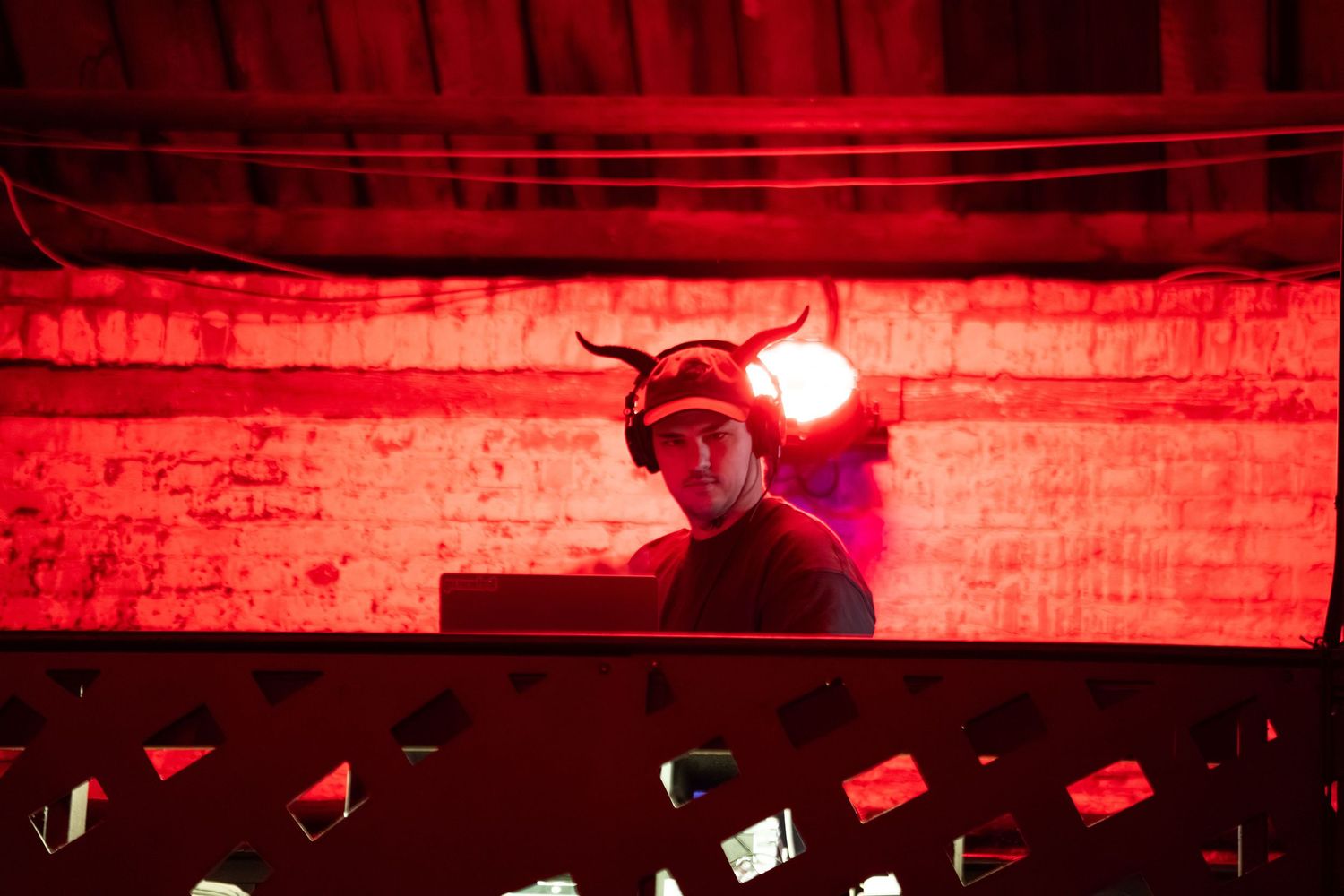 DJ performing in dark venue with red lighting and horned headpiece silhouetted against brick wall.