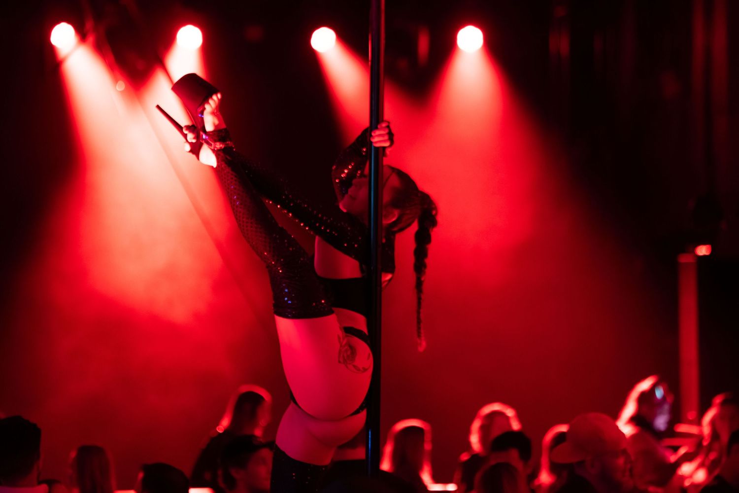 Silhouetted pole dancer performs on stage under dramatic red spotlights with audience in foreground.