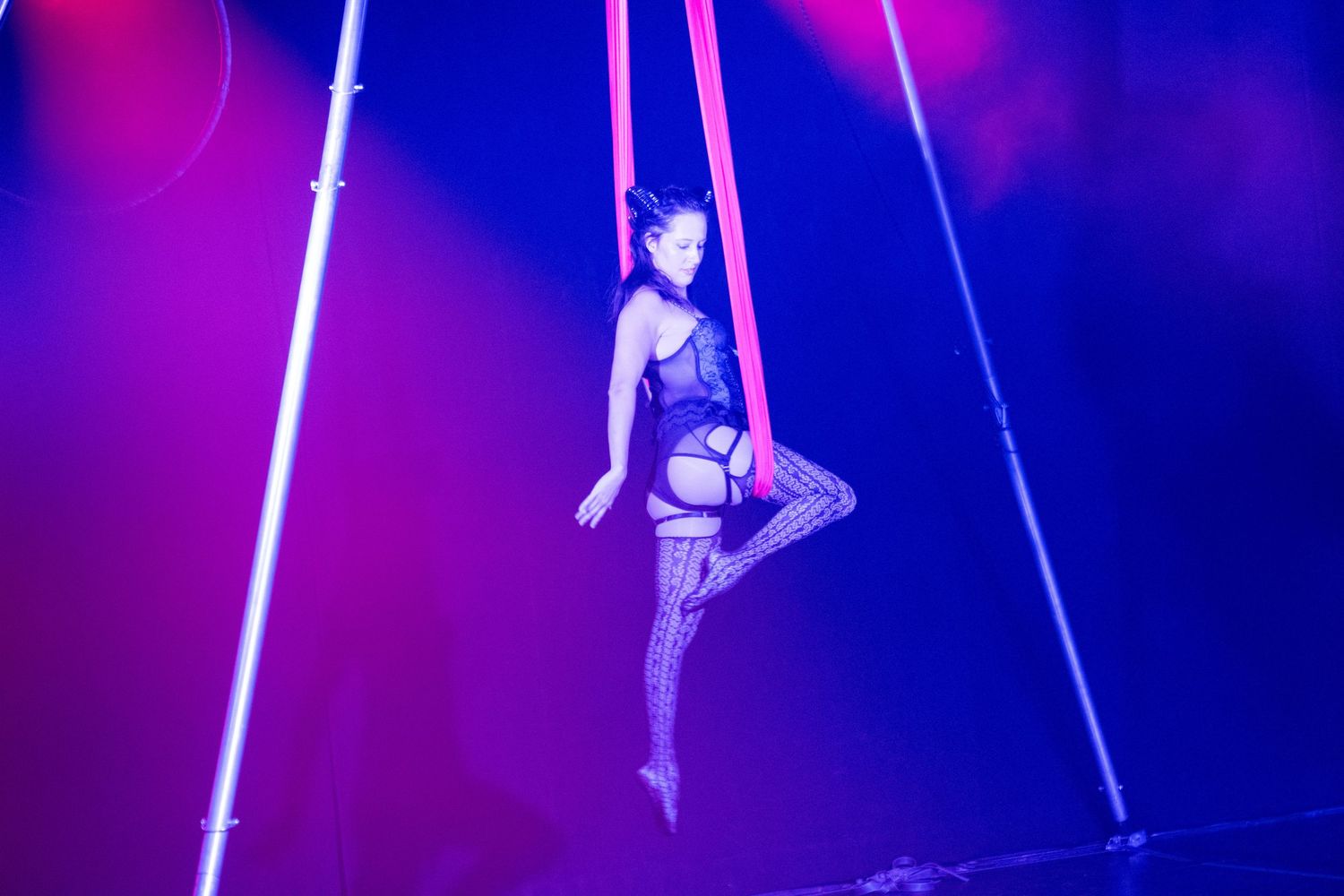 Aerial performer in purple costume gracefully poses on suspended silks against vibrant lighting.