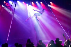 Silhouetted aerial acrobat performs against vibrant red and blue stage lighting above audience.