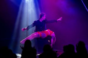 Dramatic circus performance with artist in dark costume under purple and white theatrical lighting.
