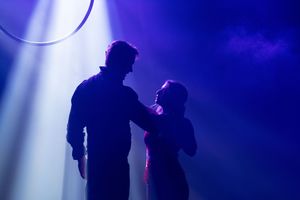 Silhouetted figures perform on stage against a dramatic purple and blue backlit backdrop.