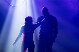 Performers in silhouette stand against a deep blue stage lighting backdrop creating an atmospheric scene.