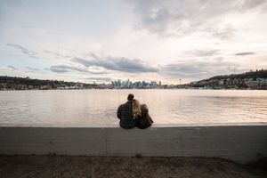 A person sits on a concrete wall overlooking Lake Union with the Seattle skyline visible in the distance at sunset.