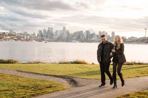 Couple walking along waterfront path with Seattle skyline view across Elliott Bay at sunset.