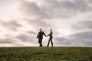 Two people holding hands while walking along a hilltop silhouetted against dramatic cloudy sunset sky.