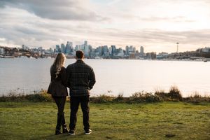 Silhouetted couple walks along grassy hilltop with Seattle skyline visible in distance during sunset.