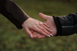 Close-up shot of hands with engagement rings in a romantic outdoor setting against a blurred green background.