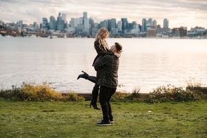 Two people embrace on a scenic overlook with Seattle's downtown skyline visible across the water at dusk.