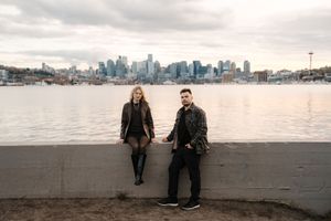 A romantic couple strolls along a misty beach at sunset with the Seattle skyline visible in the background.
