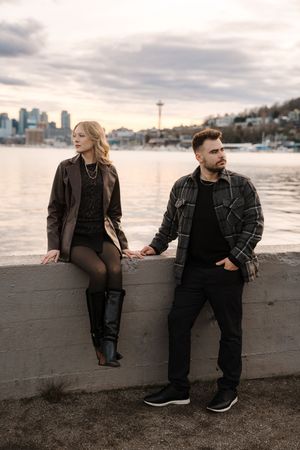 A couple stands together on a waterfront wall with the Seattle skyline and Space Needle visible in the background at dusk.