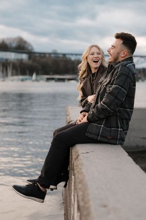 A couple laughs together while sitting on a concrete ledge by a waterfront during winter.