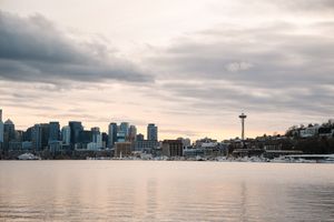 Seattle skyline at dusk with Space Needle and waterfront reflecting on calm Elliott Bay waters under cloudy skies.
