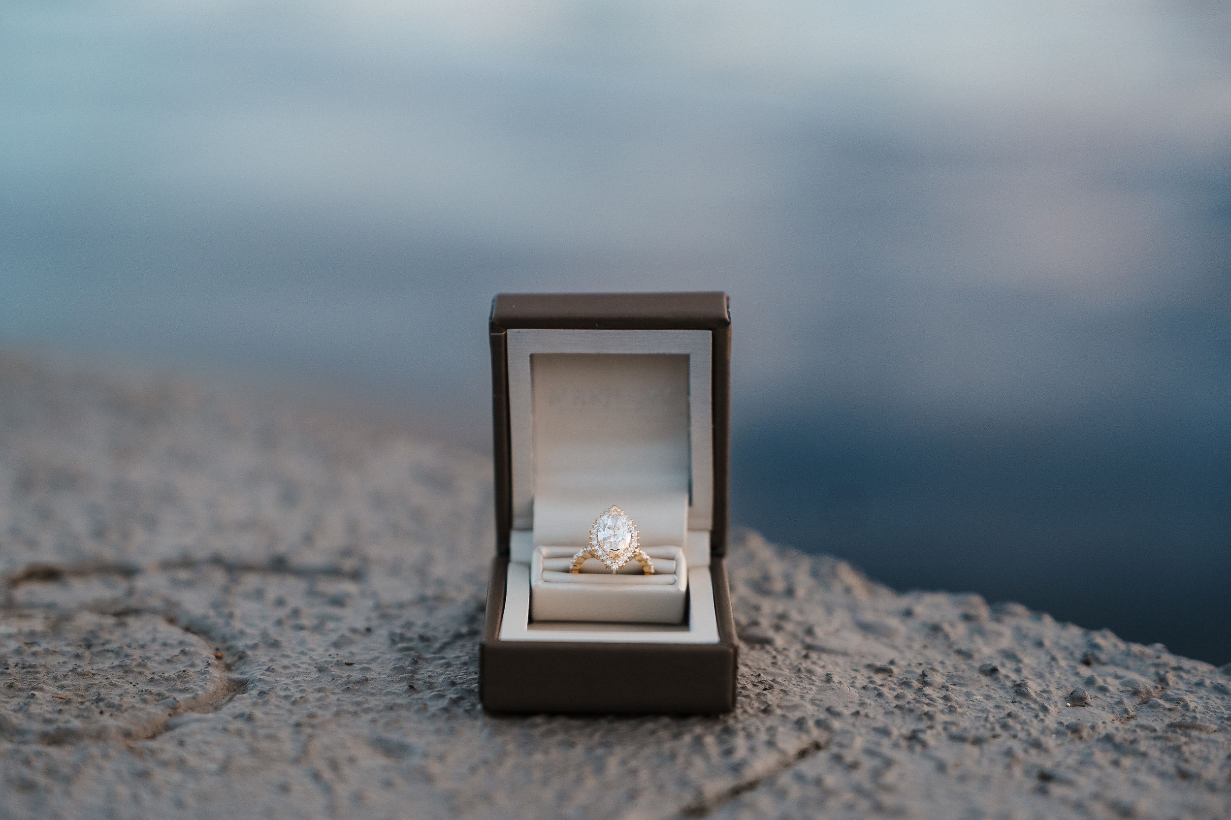 Small brown jewelry box with engagement ring sits on rocky beach terrain against moody ocean backdrop.