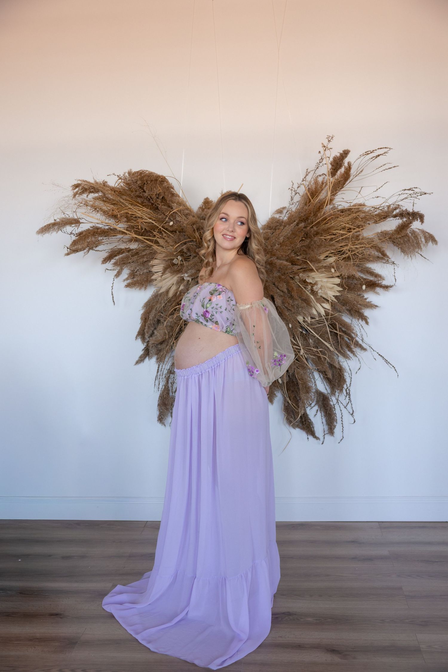 Woman in flowing lavender dress poses dynamically with dried pampas grass decorations against white backdrop.