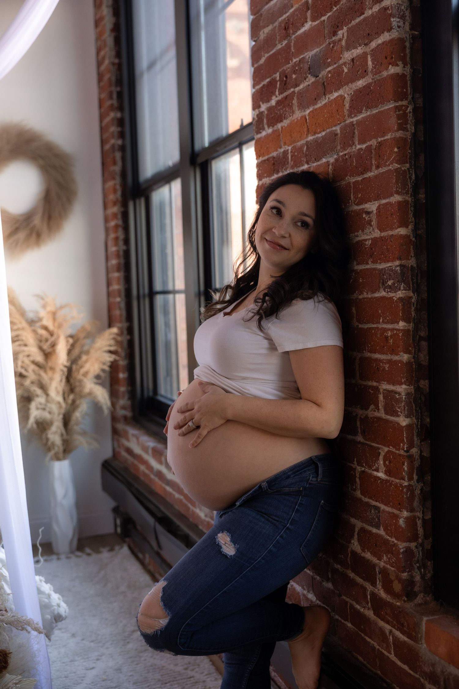 Maternity photoshoot in a rustic brick loft with natural window light and casual styling.