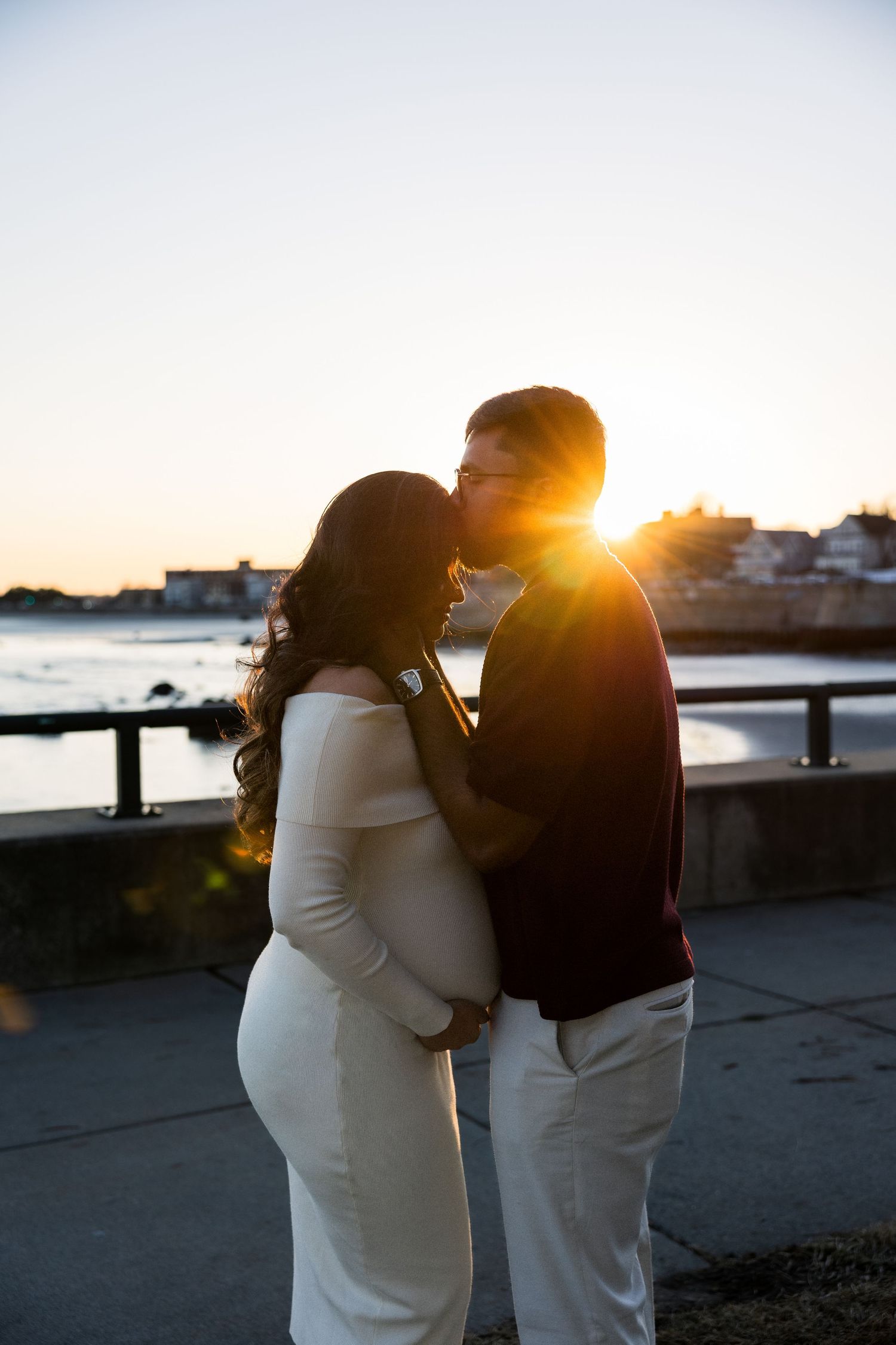 A couple in white outfits embraces while watching a golden sunset over the water at a seaside location.