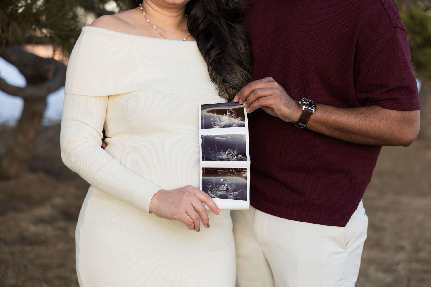 A couple in wedding attire holds ultrasound photos while embracing outdoors in autumn.