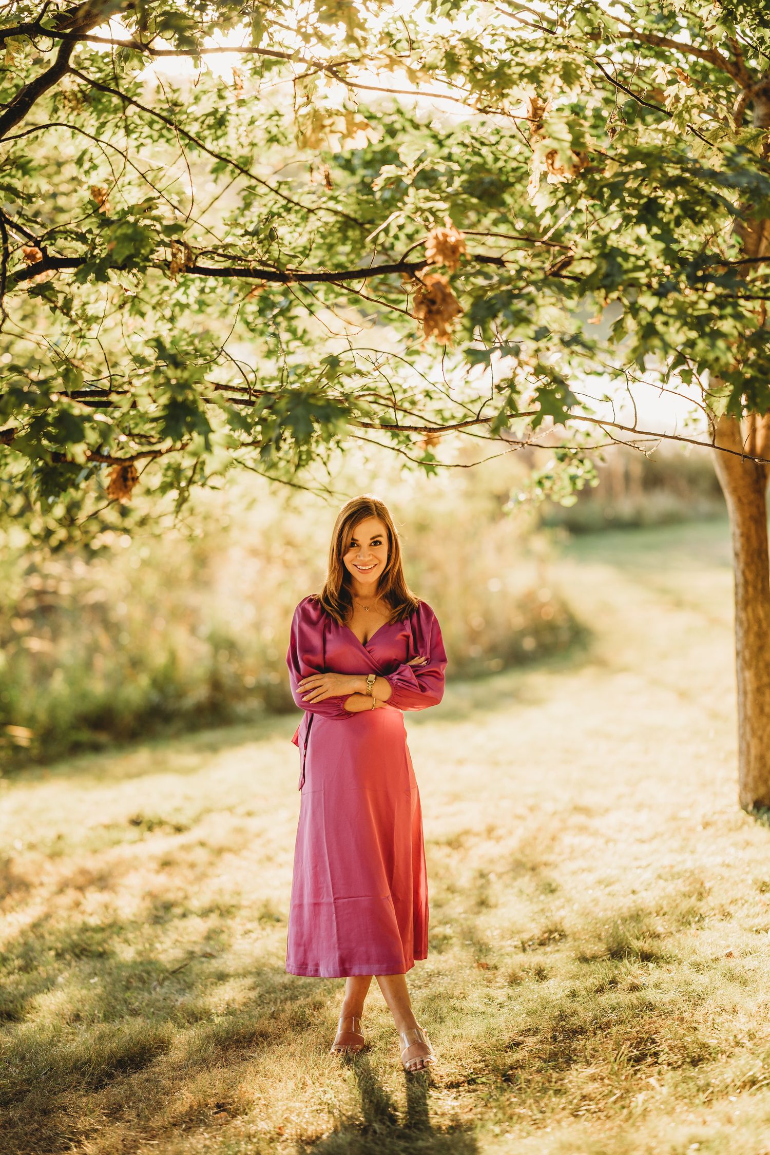 A person in a burgundy dress stands under a sunlit tree in a dreamy outdoor setting during golden hour.