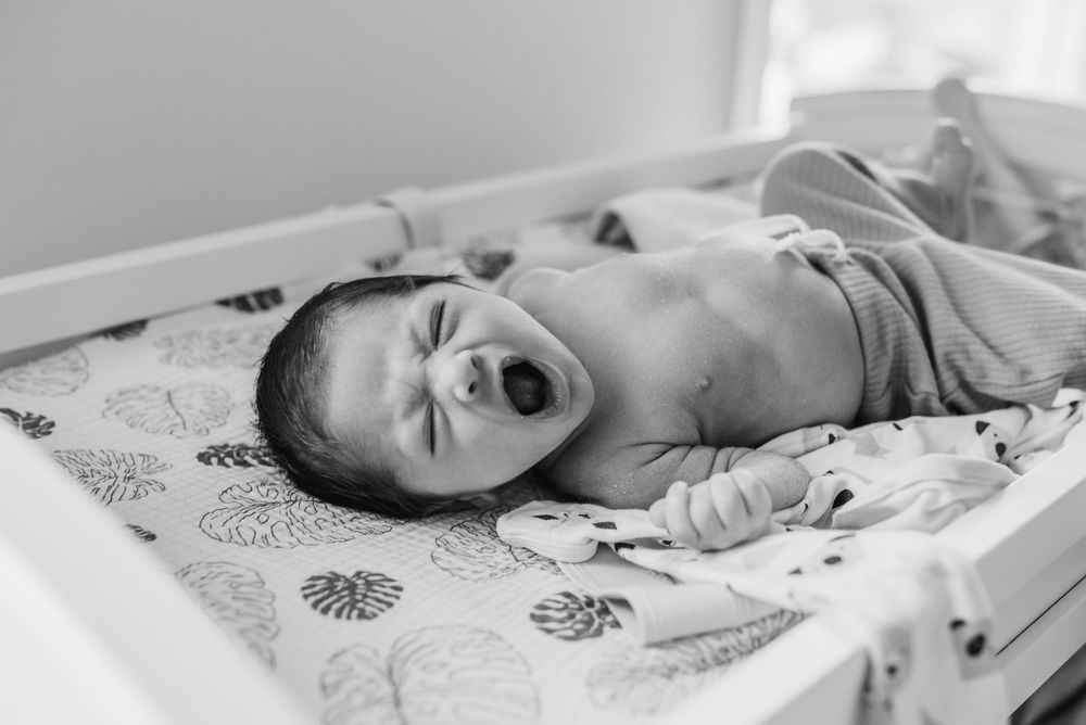 Black and white photo of a baby lying on a patterned blanket while yawning or crying.