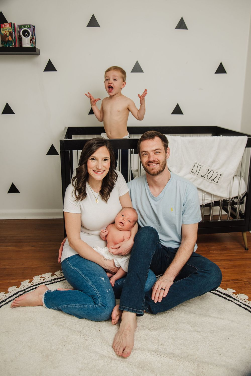 Family sitting together on white rug in nursery with black triangle wall decals and wooden crib.