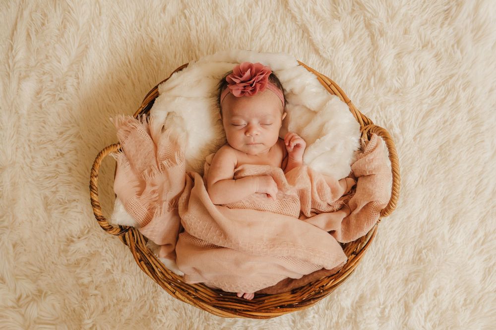Sleeping newborn baby wrapped in soft pink fabric lying in woven basket against cream colored textured backdrop.