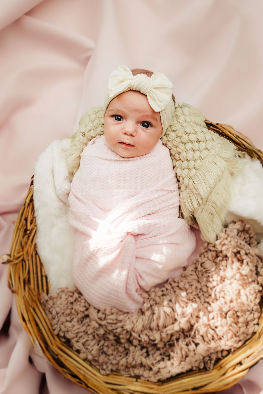Baby in pink swaddle and white bow headband nestled in wicker basket with cozy textured blanket.