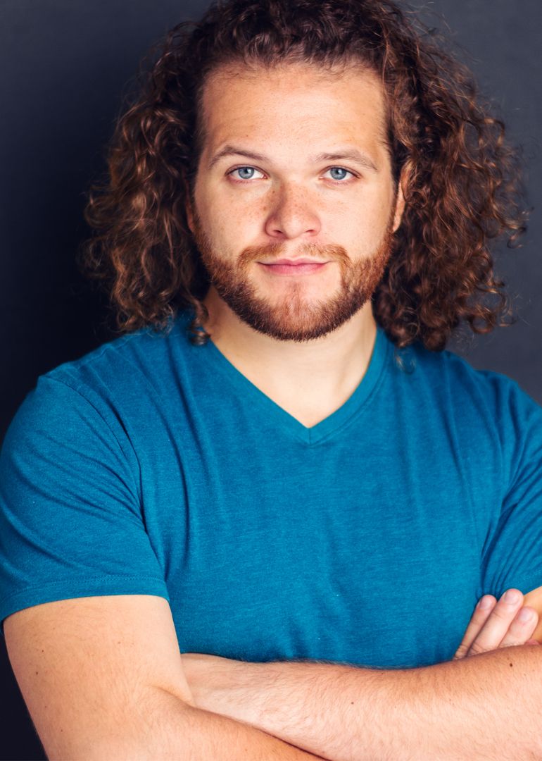 A person with curly hair and a beard wearing a teal v-neck shirt poses against a dark background in a headshot series.