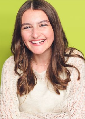 Smiling person with long brown hair wearing a cream colored knit sweater against a bright lime green background.