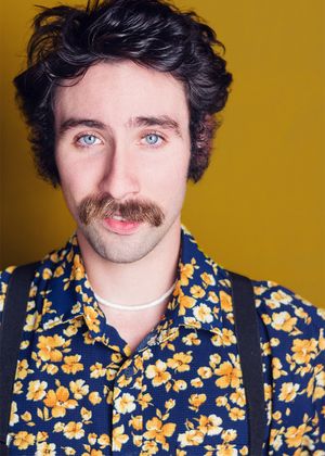 Person wearing floral print shirt and black suspenders poses against golden background.