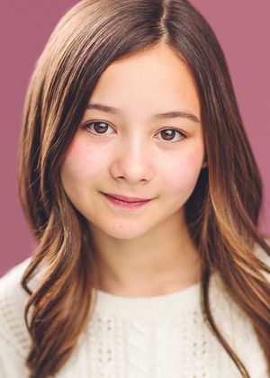 Young girl with long brown hair and brown eyes smiling softly, wearing a white sweater against a pink background.