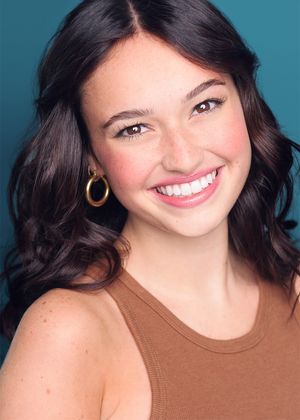 Professional headshot of a smiling young woman with dark wavy hair wearing a brown tank top and gold hoop earrings.