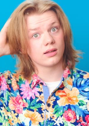 Portrait against bright blue background wearing vibrant floral Hawaiian shirt with pink, orange and yellow tropical flowers.