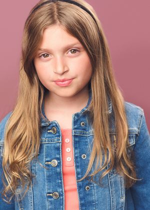 Young person with long blonde hair wearing a denim jacket and coral top against a pink background.