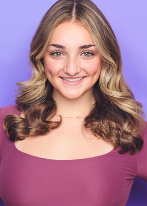 Portrait of a smiling person with wavy blonde and brown hair wearing a burgundy top against a purple background.