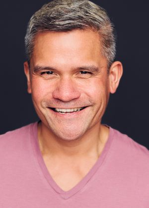Portrait of a middle-aged person in a pink v-neck shirt smiling warmly against a dark background.