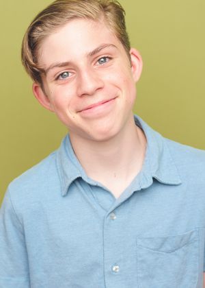 Professional headshot of a young person with blonde hair wearing a light blue collared shirt against a green background.