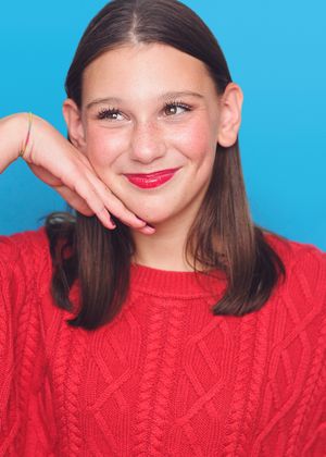 Young person in a red cable knit sweater poses playfully with hand on chin against bright blue background.