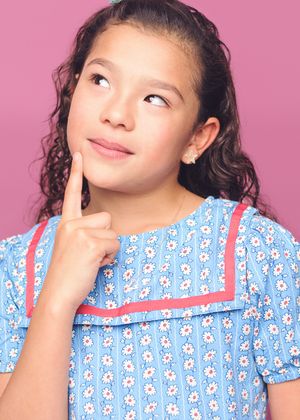 Young person in floral print dress poses thoughtfully with finger on chin against pink background.