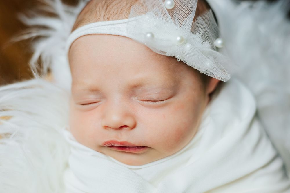 Sleeping newborn baby wearing white cap and lace details in soft lighting.