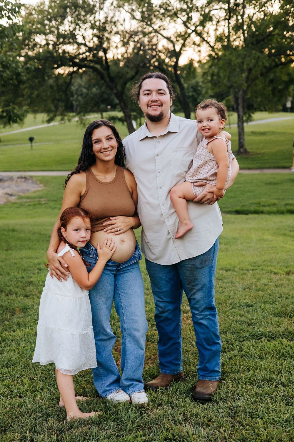 A happy family of four poses outdoors on green grass, with a pregnant mother, father, and two young daughters at golden hour.