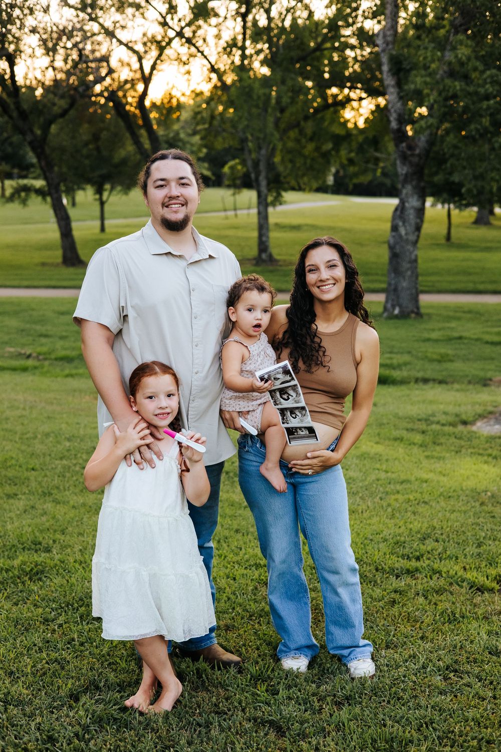 Family of four poses outdoors at sunset, mom holding ultrasound photos, announcing a new pregnancy.