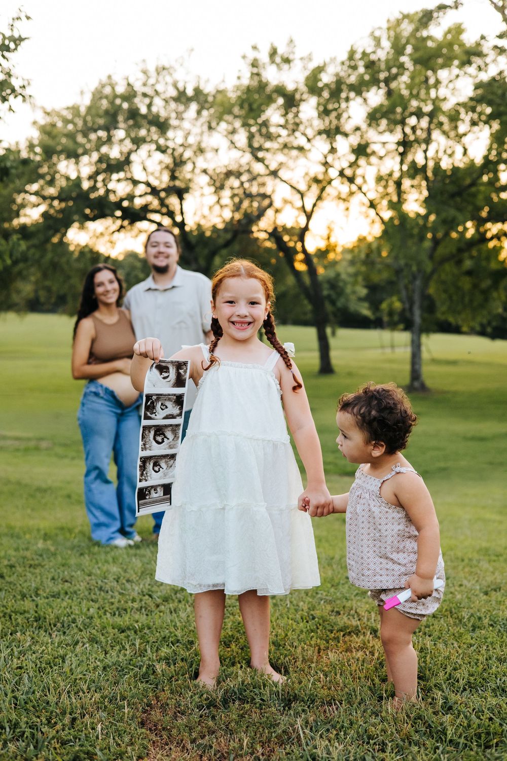 Family announces pregnancy outdoors at sunset, girl in white dress holds ultrasound photos with toddler sister.