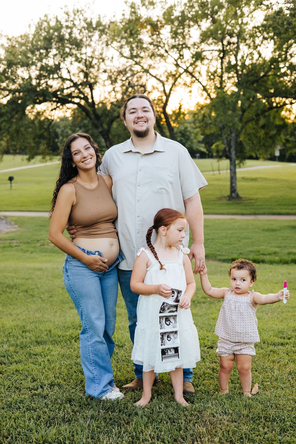 Pregnant woman and man pose with two young daughters in a sunny park, older girl holds ultrasound photos.
