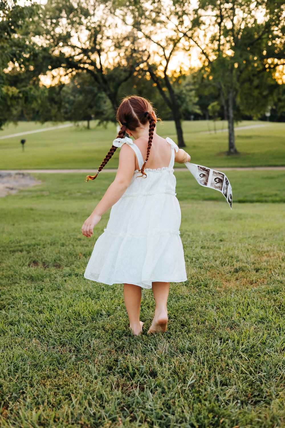 Young girl with red braided pigtails in white dress runs barefoot through green park at golden hour sunset.