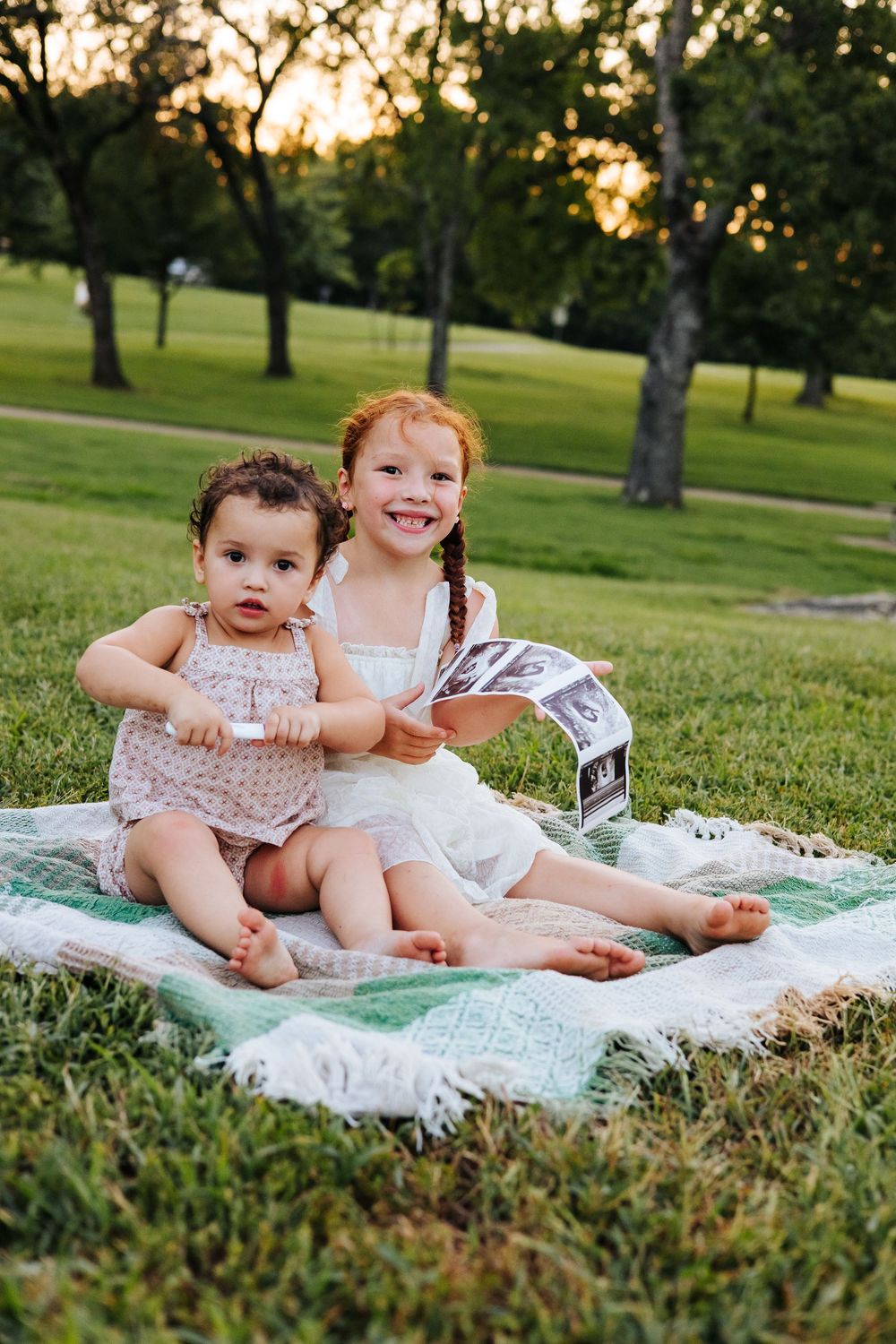 Two young girls sit on a blanket in a park, smiling and holding ultrasound photos at golden hour.