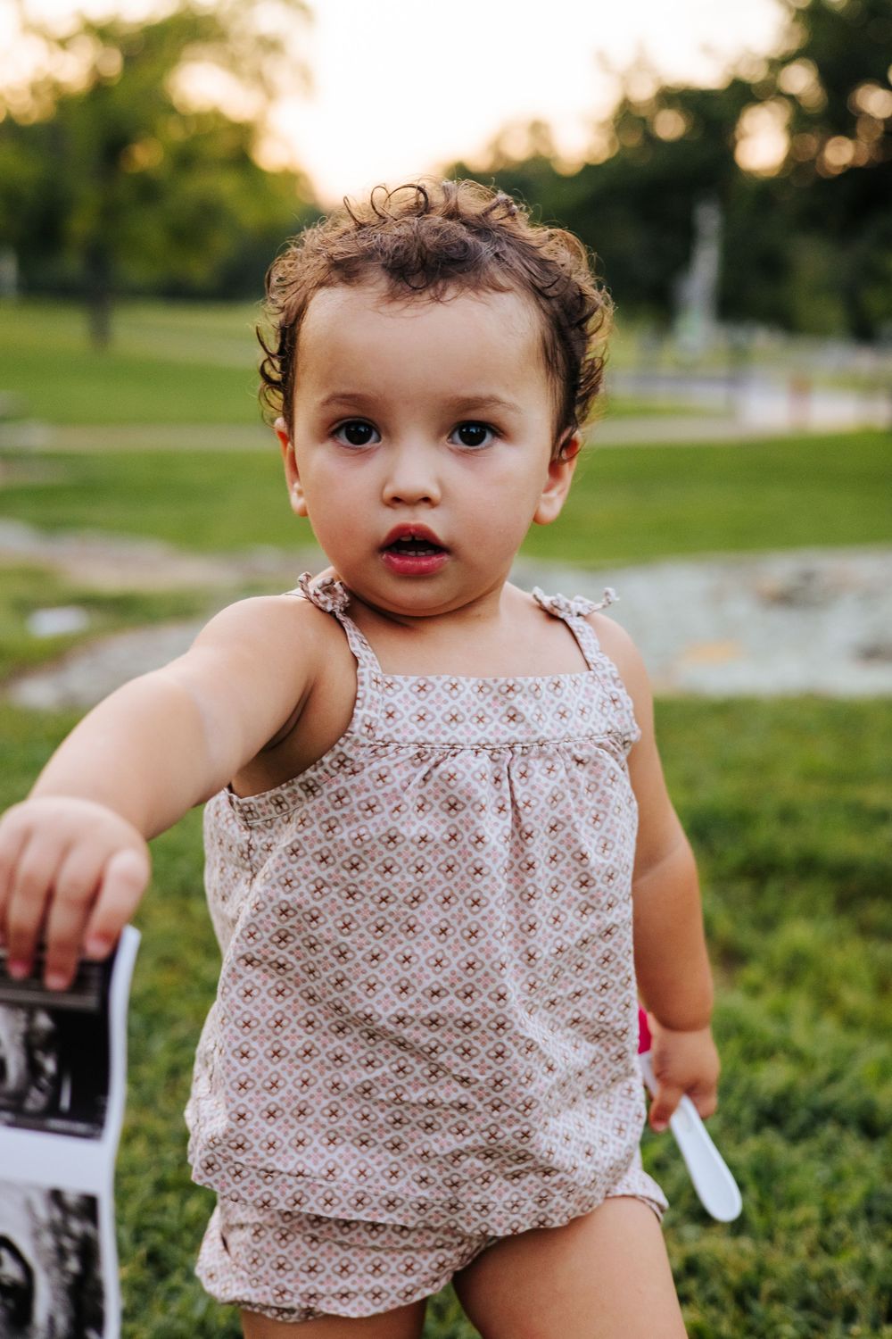 Curly-haired toddler girl in floral romper holding ultrasound photos while standing on green grass outdoors.