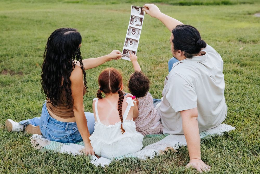 Family sitting on grass holding ultrasound photos, announcing pregnancy with two young children during outdoor picnic.